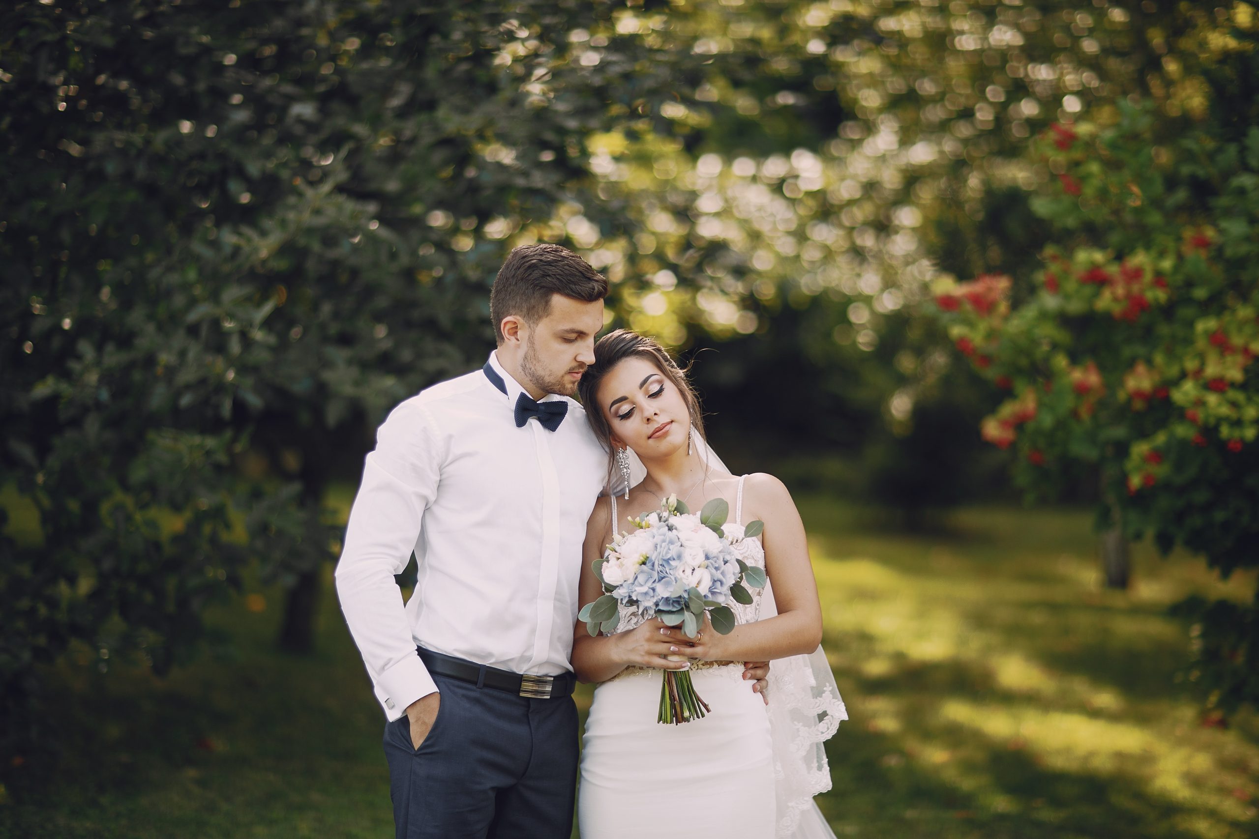 A young and beautiful bride and her husband is standing in a summer park with bouquet of flowers